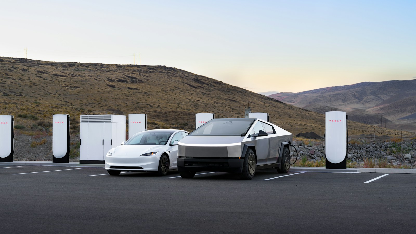 White Tesla Model 3 and silver Tesla Cybertruck parked at a charging station with multiple Tesla chargers in a mountainous area at dusk.